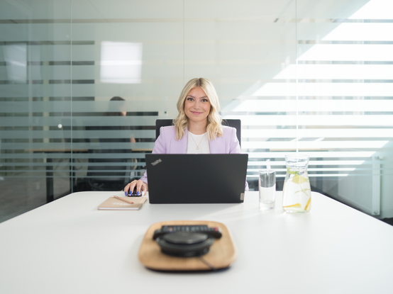 Una persona è seduta a un tavolo in una sala riunioni moderna, utilizzando un laptop. Sul tavolo ci sono un bicchiere d'acqua, una caraffa con limone e un taccuino. La stanza è illuminata da luce naturale e presenta pareti in vetro con strisce orizzontali.