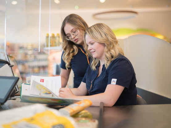 Due persone stanno discutendo e consultando documenti in un negozio. Sul tavolo ci sono fogli e un dispositivo elettronico. Sullo sfondo si intravedono prodotti esposti in un negozio.