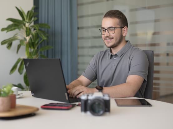 Una persona sta lavorando al computer in un ambiente moderno e luminoso. Sul tavolo ci sono un laptop, un tablet, un telefono e una macchina fotografica. Sullo sfondo si intravede una pianta e una tenda.