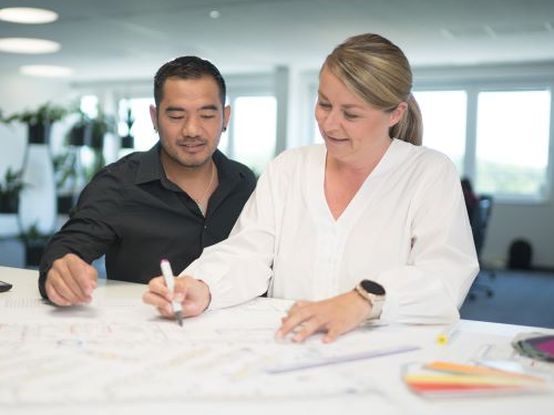 Due persone collaborano su un progetto, esaminando documenti e piani su un tavolo in un ambiente di lavoro luminoso e moderno.