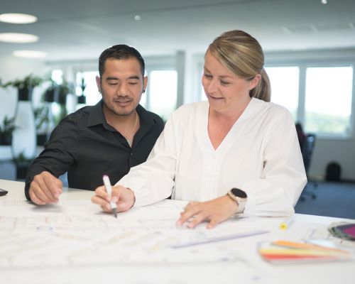 Due persone collaborano su un progetto, esaminando documenti e piani su un tavolo in un ambiente di lavoro luminoso e moderno.