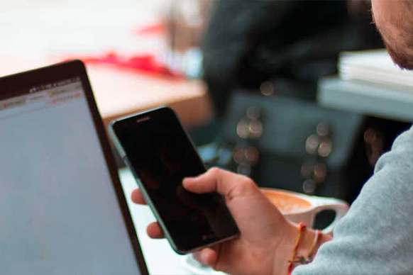 Un uomo tiene in mano uno smartphone mentre si trova accanto a un laptop e una tazza di caffè.