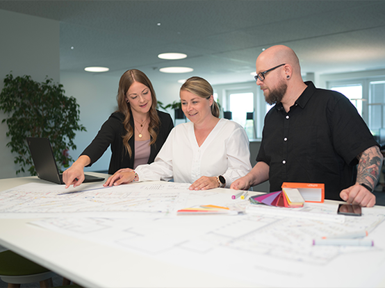 Un gruppo di tre persone discute su un tavolo, esaminando dei piani o progetti. L'ambiente è luminoso e moderno, con piante in background.
