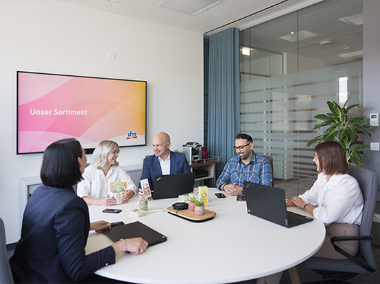 Un gruppo di persone partecipa a una riunione in una sala conferenze, con una presentazione visibile su uno schermo e vari materiali di lavoro sul tavolo.